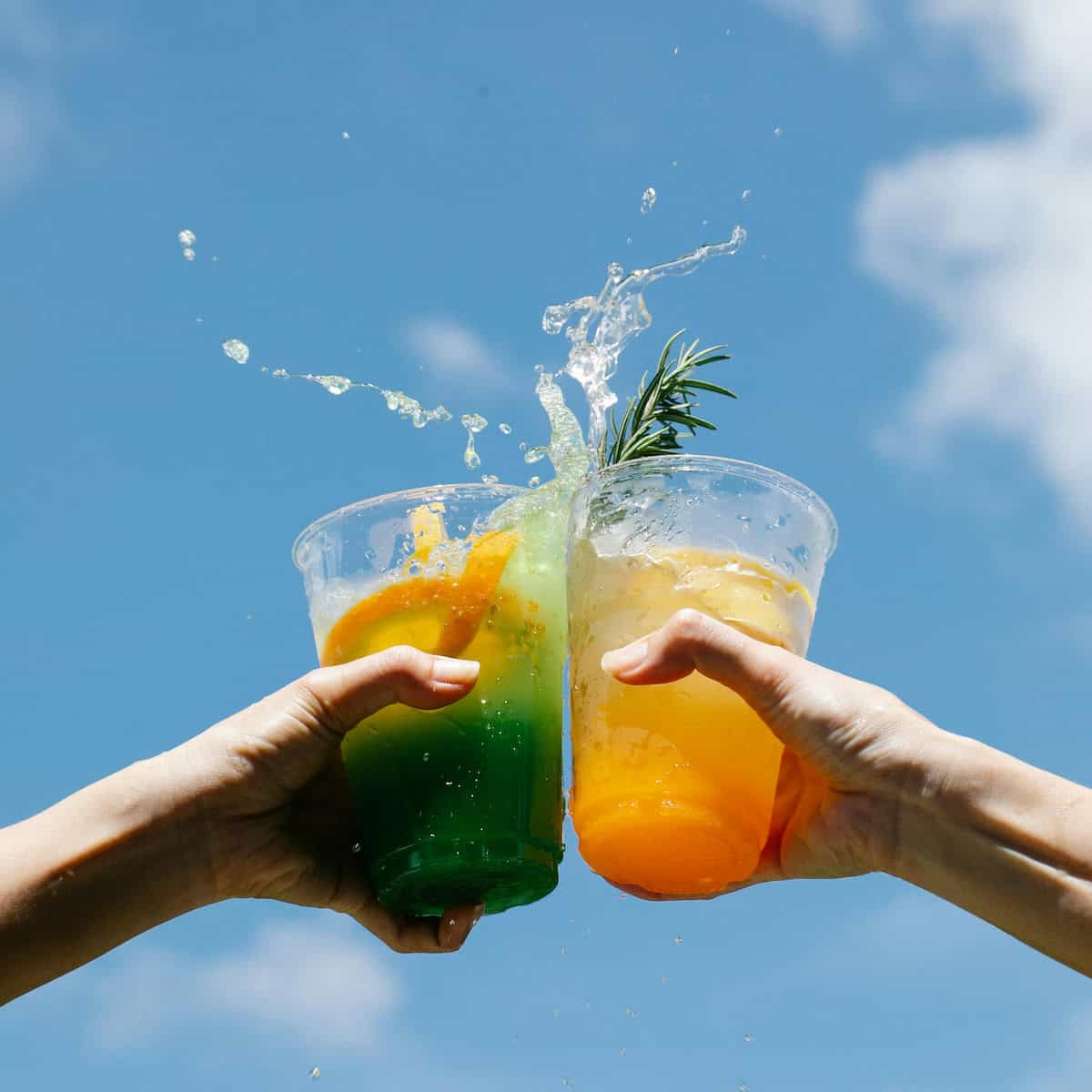 Two hands holding plastic cups of vibrant Jamaican drinks in mid-air toast, with liquid splashing against a blue sky backdrop.