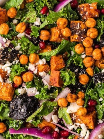 A bowl of kale salad topped with roasted chickpeas, sweet potatoes, feta cheese, red onions, dried fruit, and pomegranate seeds on a wooden surface.