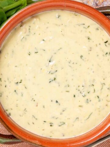 A bowl of creamy soup with herbs, placed on a brown cloth next to a metal spoon and fresh basil leaves.