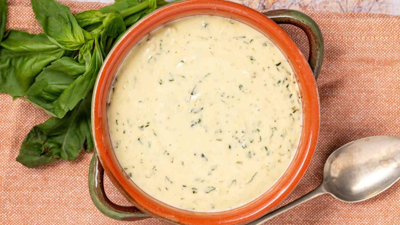 A bowl of creamy soup with herbs, placed on a brown cloth next to a metal spoon and fresh basil leaves.