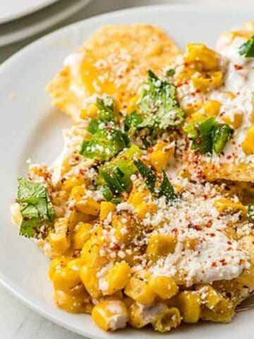 A white plate with tortilla chips topped with corn, cilantro, cheese, crema, and chili powder. A lime wedge is visible nearby.
