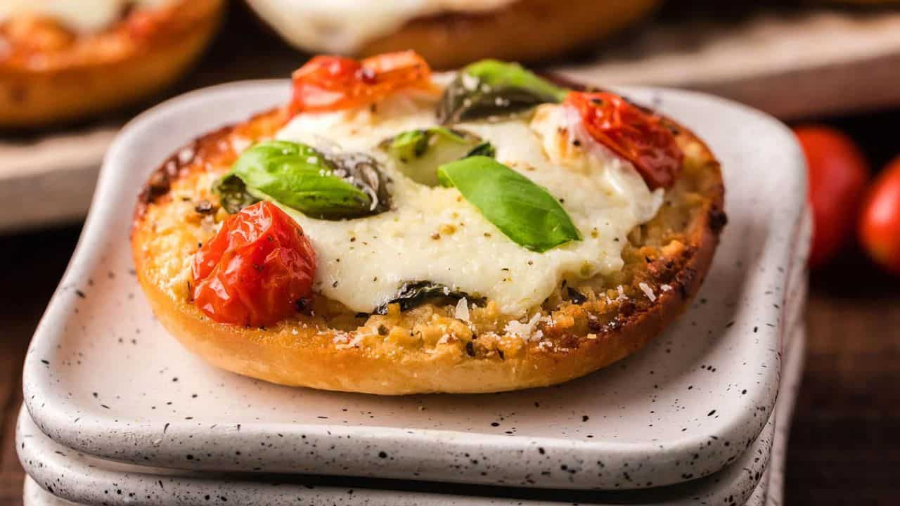 A toasted bagel half topped with melted mozzarella cheese, roasted tomatoes, basil leaves, and black pepper on a stack of small square plates.
