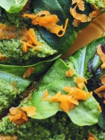 Three triangular slices of flatbread topped with green pesto, fresh spinach leaves, and shredded carrots on a yellow plate.