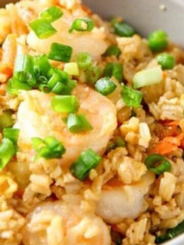 A bowl of shrimp fried rice garnished with chopped green onions, containing peas, carrots, and rice, with chopsticks beside the bowl.