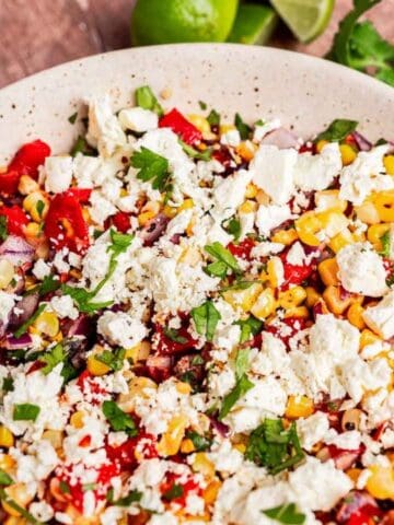 A bowl of salad with corn, diced red peppers, onions, chopped herbs, and crumbled white cheese, with a serving spoon and lime wedges in the background.