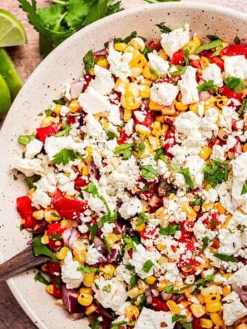 A bowl of salad with corn, tomatoes, red onion, chopped herbs, and crumbled cheese sits on a wooden surface next to fresh limes and cilantro.