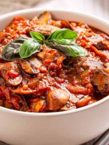 A white bowl filled with mushroom and tomato stew, garnished with fresh basil, sits next to a spoon on a light surface.
