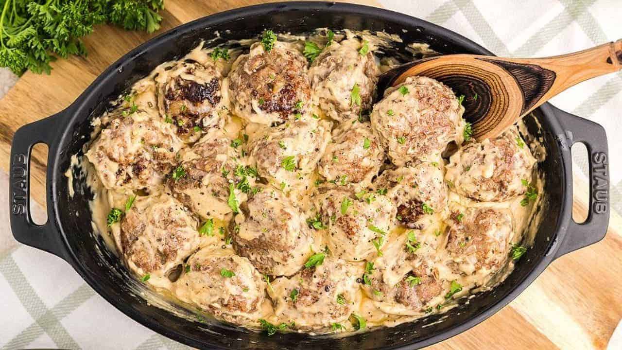 A black oval dish filled with meatballs covered in creamy sauce, garnished with chopped herbs, with a wooden spoon resting on the side.