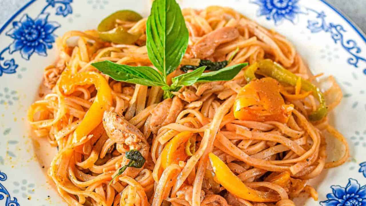 Plate of stir-fried noodles with sliced chicken, yellow bell peppers, green peppers, onions, and a garnish of fresh basil on top.