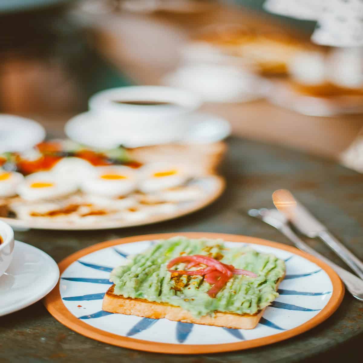 A plate with avocado toast topped with red onions in the foreground, and a blurred plate of eggs and coffee cup in the background on a table.