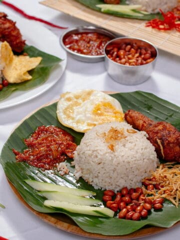 Plates of nasi lemak with rice, fried chicken, egg, peanuts, anchovies, cucumber, and sambal served on banana leaves, with extra sambal and peanuts in small bowls.