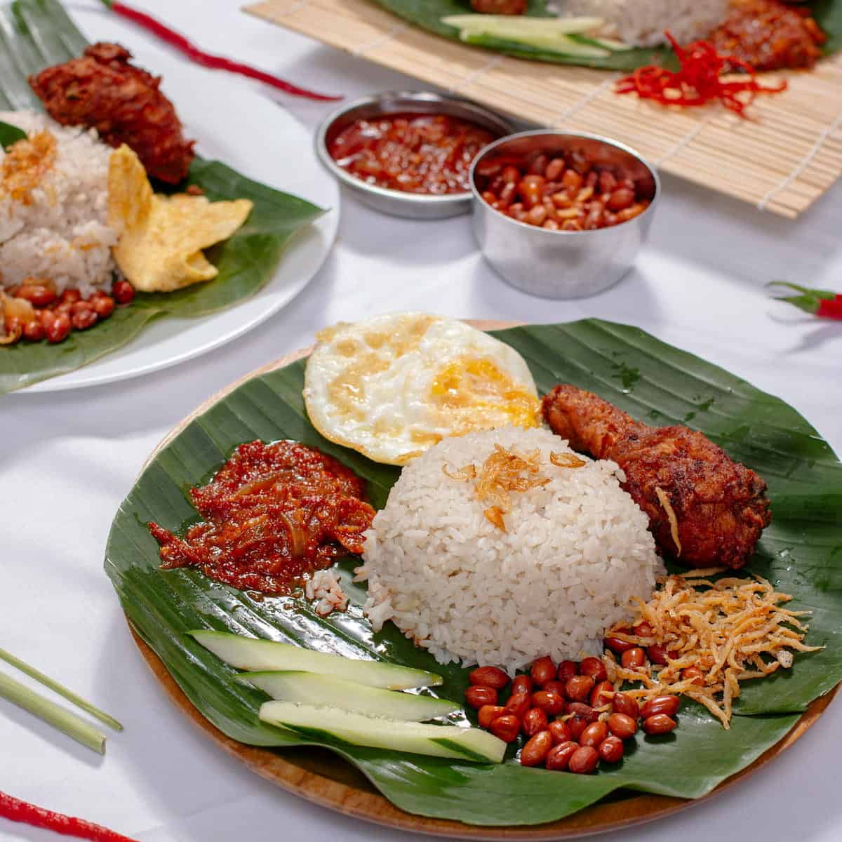 Plates of nasi lemak with rice, fried chicken, egg, peanuts, anchovies, cucumber, and sambal served on banana leaves, with extra sambal and peanuts in small bowls.