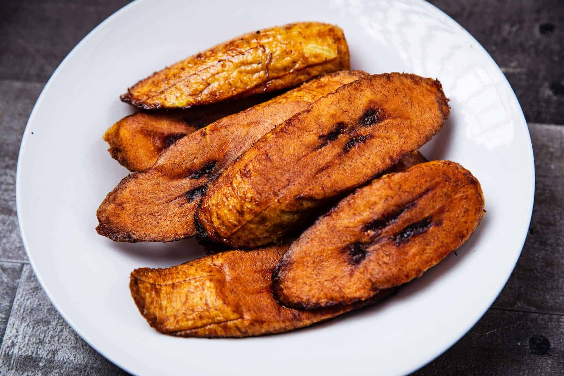 A white plate with several pieces of fried ripe plantains arranged on it, placed on a dark wooden surface.