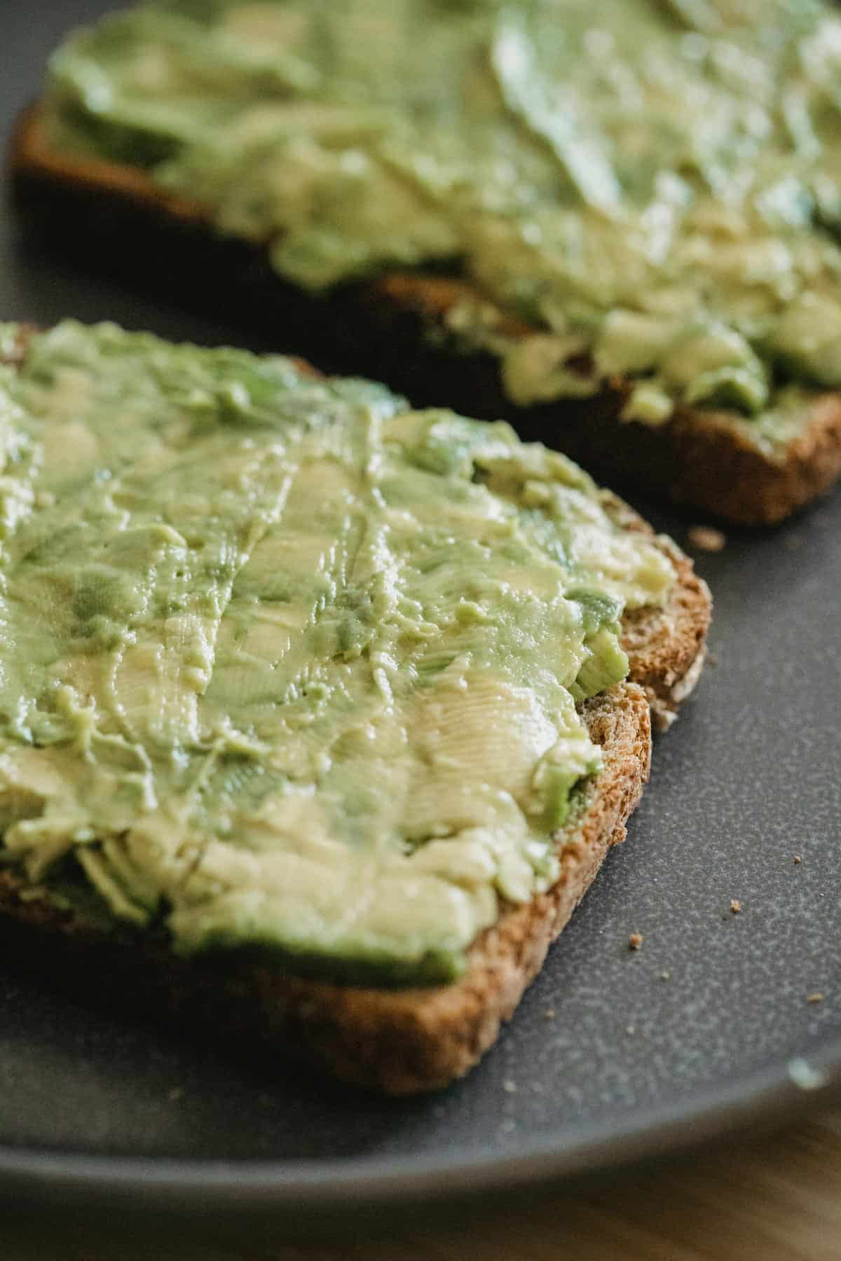 Two slices of whole grain bread topped with mashed avocado on a dark plate.
