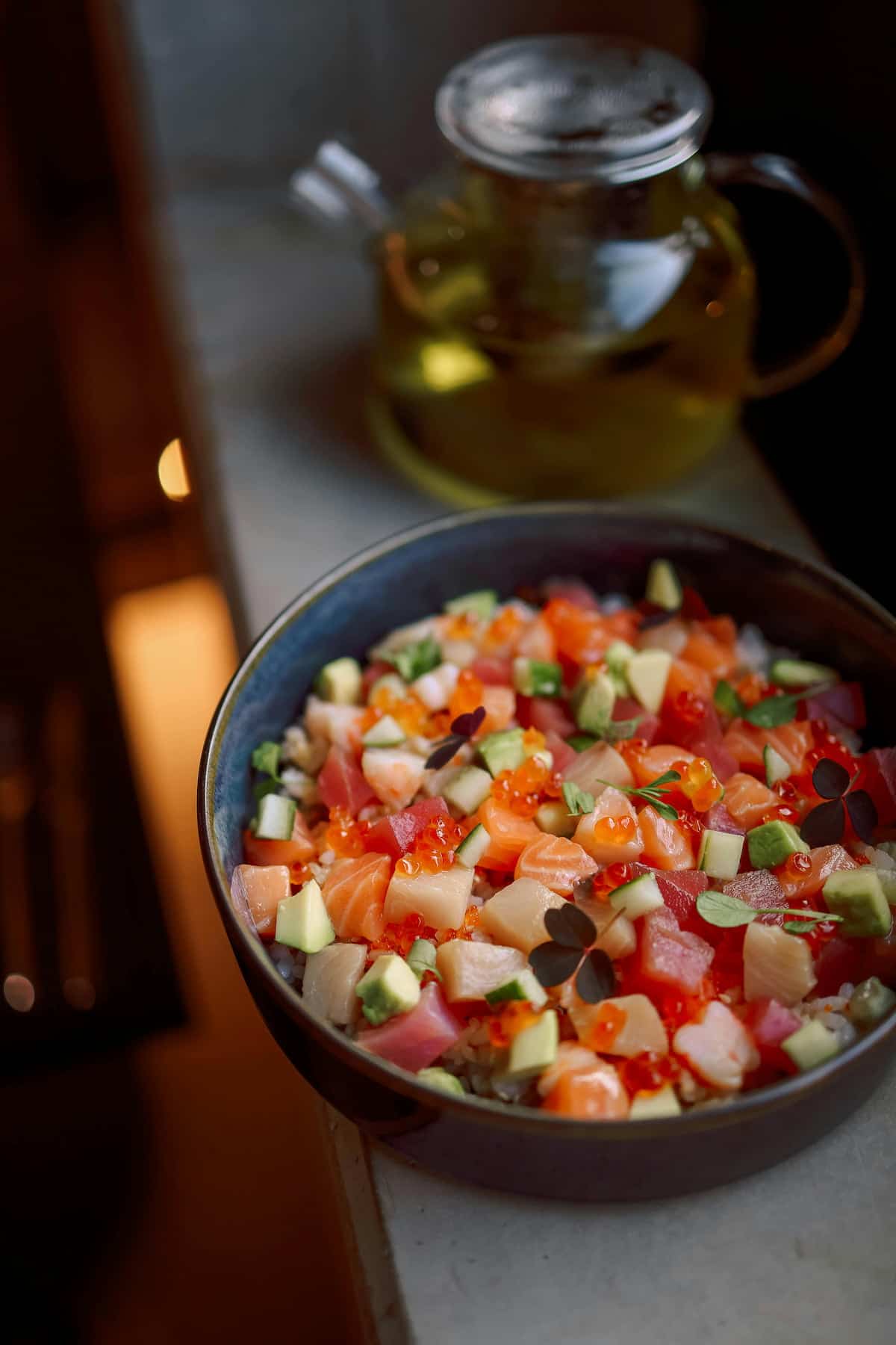 A bowl of poke featuring diced salmon, tuna, avocado, and garnishes, with a glass teapot in the background.