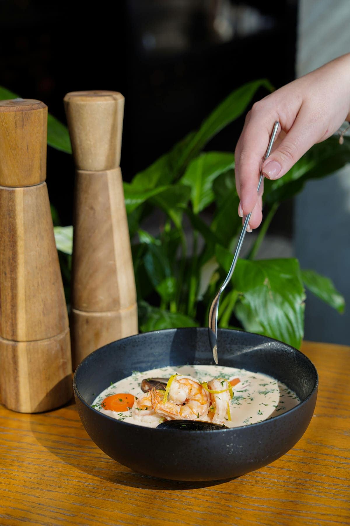 A hand holds a spoon above a black bowl of creamy seafood soup with shrimp and vegetables, set on a wooden table beside two wooden pepper mills and green plants.