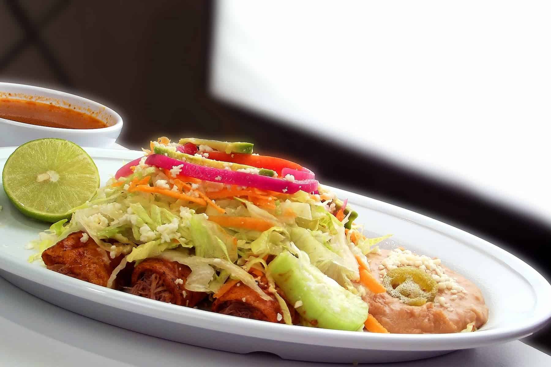 A plate of enchiladas topped with lettuce, pickled onions, and cheese, served with refried beans, a lime wedge, and a bowl of red sauce.