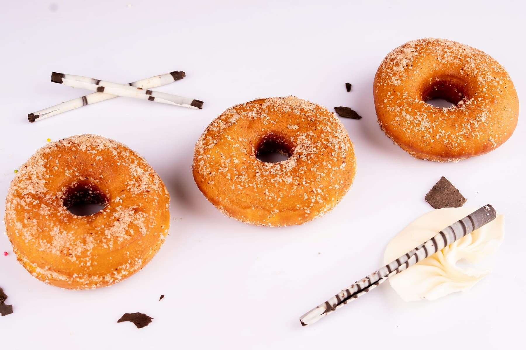 Three sugar-coated donuts are arranged on a white surface with chocolate sticks, chocolate pieces, and a swirl of white cream.