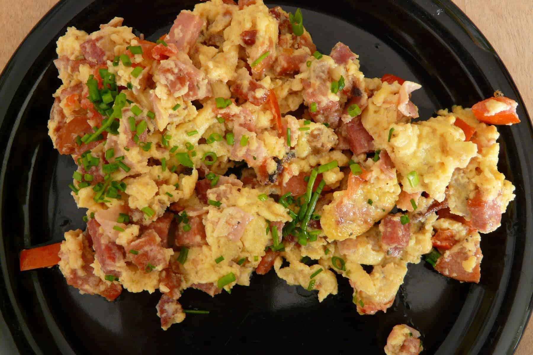 Scrambled eggs mixed with chopped ham, tomatoes, and green herbs, served on a black plate.
