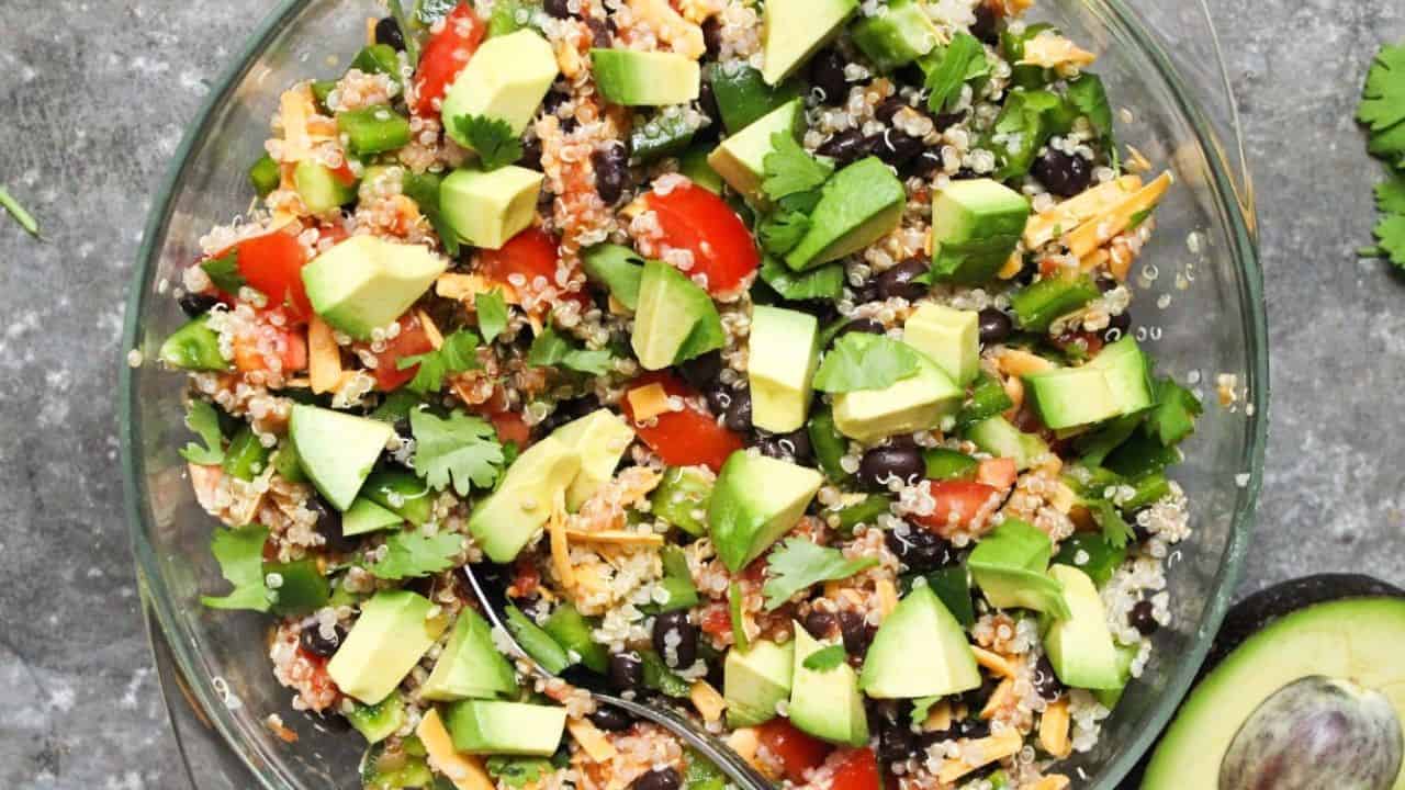 Quinoa taco salad in a large glass bowl with diced avocado and shredded cheese on top, next to a small bowl of salsa.