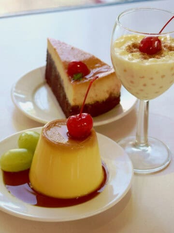 A plate with flan topped with a cherry and grapes, a plate with a slice of flan cake, and a glass of tapioca pudding topped with a cherry, all on a white surface.