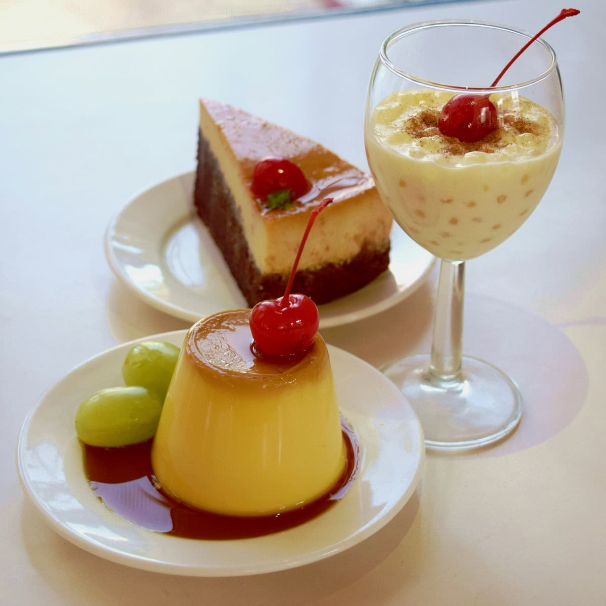 A plate with flan topped with a cherry and grapes, a plate with a slice of flan cake, and a glass of tapioca pudding topped with a cherry, all on a white surface.