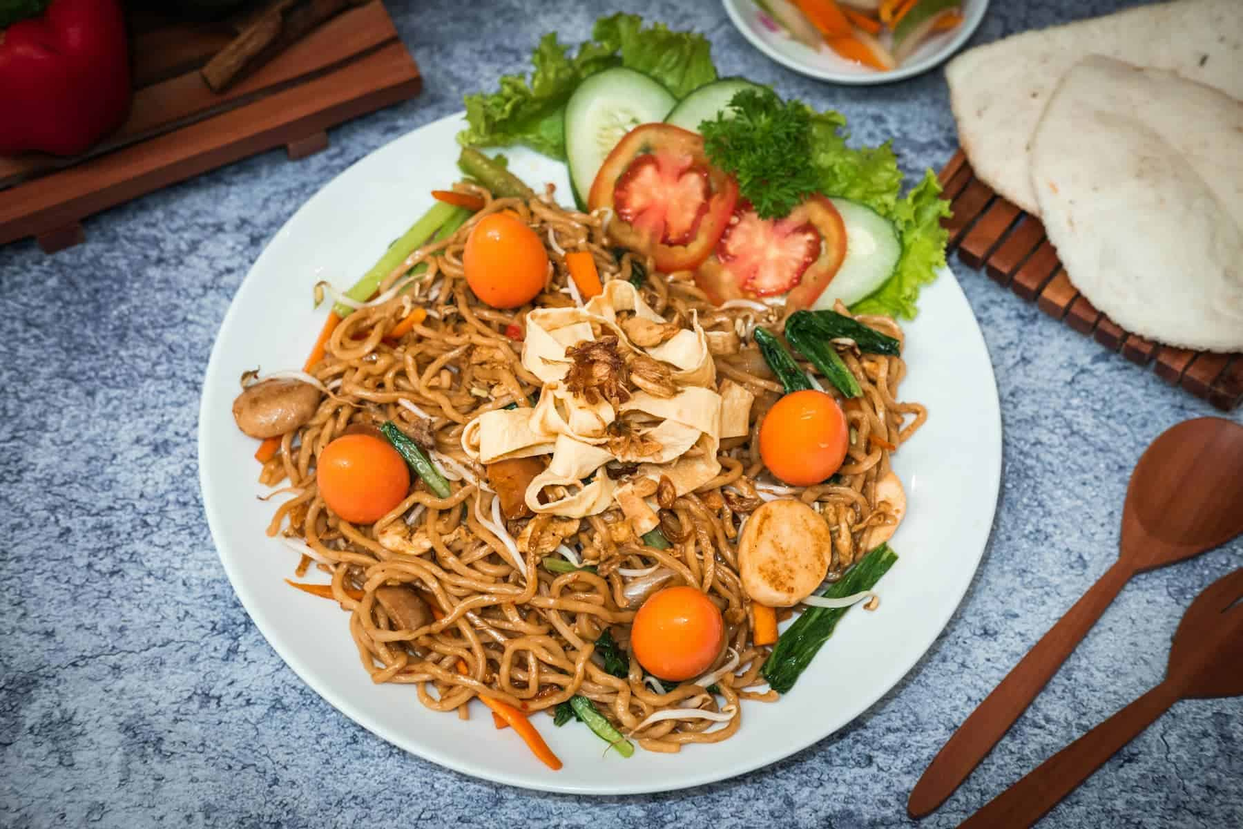 A plate of stir-fried noodles with vegetables, quail eggs, and seafood, garnished with cucumber, lettuce, and tomato slices. Wooden cutlery and flatbreads are visible beside the plate.