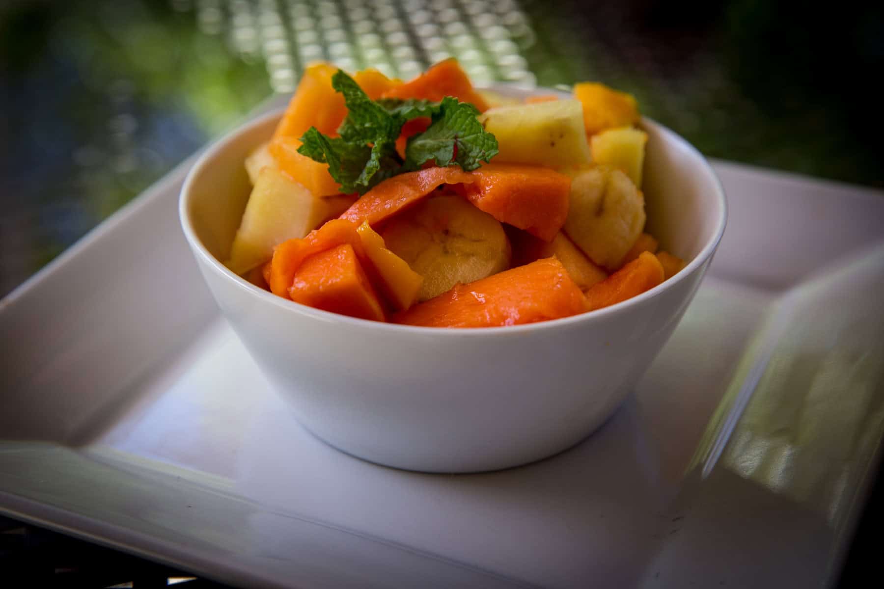 A white bowl filled with cut pieces of papaya, banana, and pineapple, garnished with a sprig of mint, sits on a white plate.