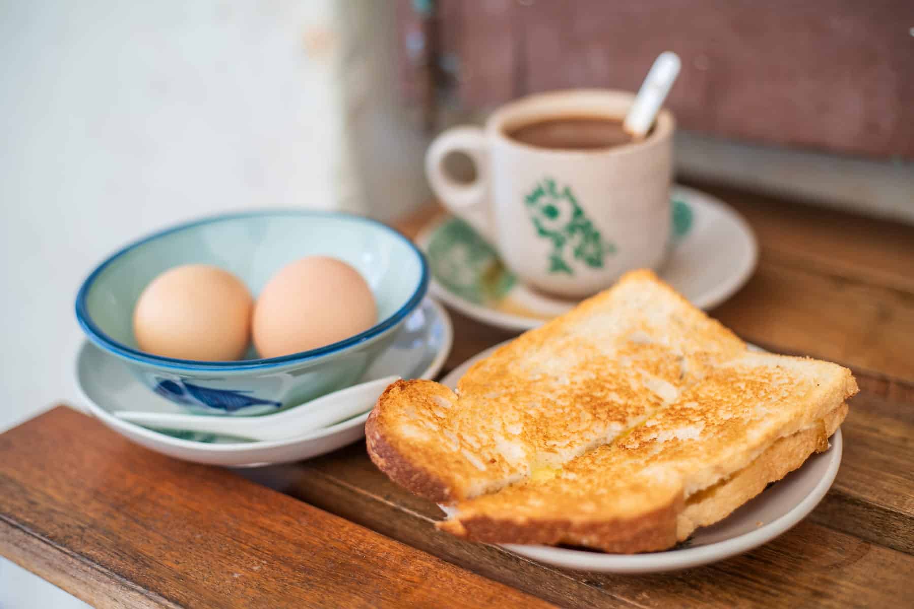 A plate of toasted bread, a bowl with two eggs, and a cup of coffee on a wooden table.
