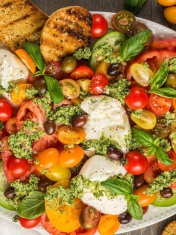 A platter of colorful sliced tomatoes, mozzarella, olives, basil, and pesto, garnished with grilled bread slices.