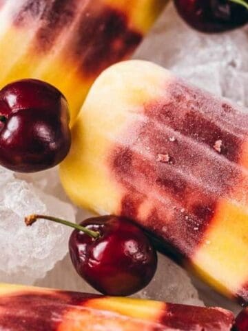 Assorted cherry and yellow popsicles with fresh cherries are arranged on ice.