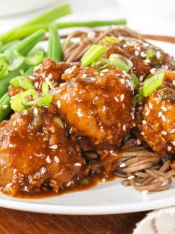 A plate of saucy meatballs on noodles, garnished with sesame seeds and chopped green onions, with green beans on the side.