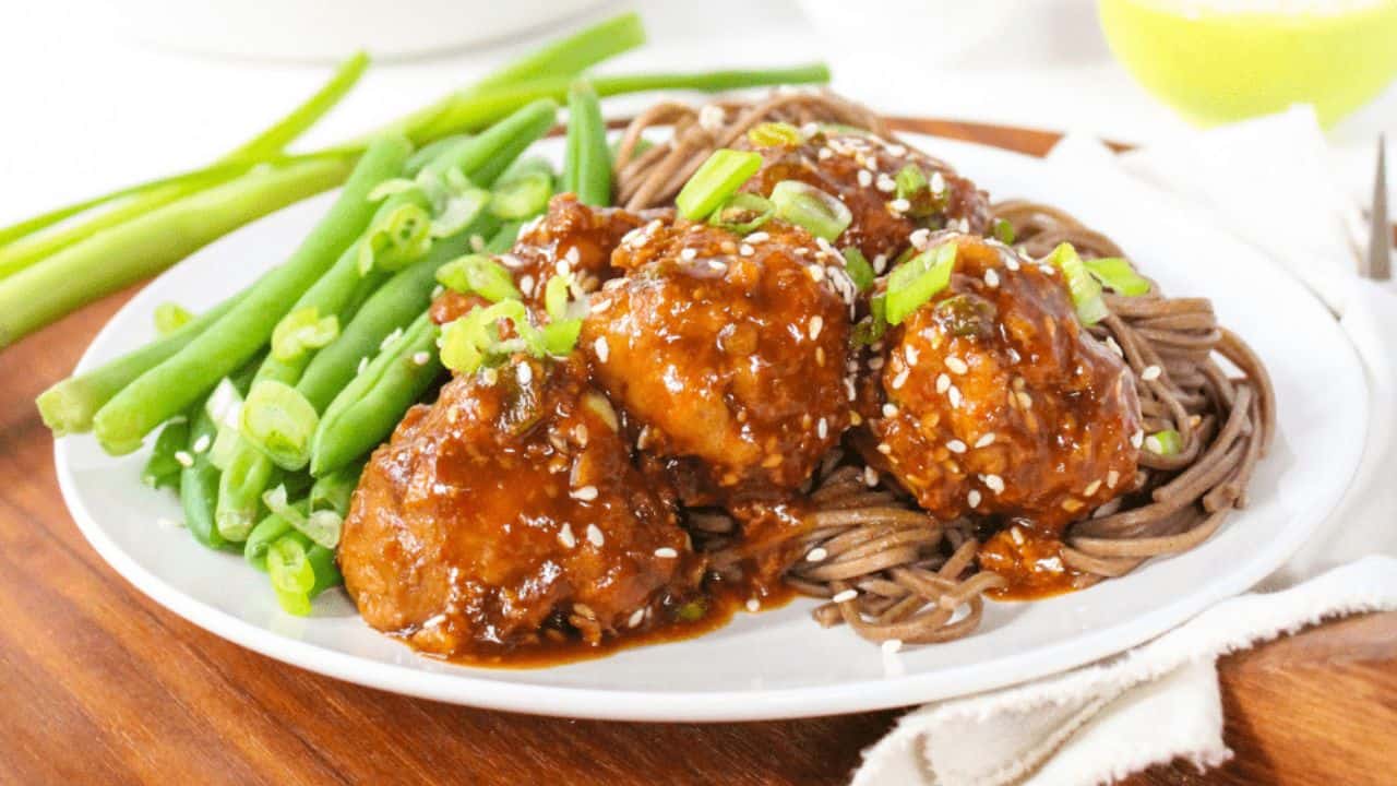 A plate of saucy meatballs on noodles, garnished with sesame seeds and chopped green onions, with green beans on the side.