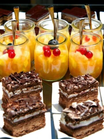Glass jars with mixed fruit salad topped with berries and metal spoons, displayed behind chocolate layered cake squares on white napkins.