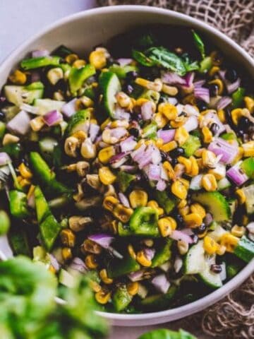 A bowl of salad containing corn, black beans, cucumbers, red onions, spinach, and chopped herbs, with a fork resting inside the bowl.