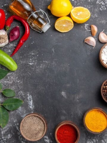 Assorted vegetables, spices, herbs, lemon halves, garlic, and bottles of oil are arranged on a dark, textured surface.