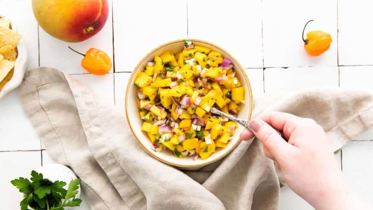 Overhead shot of a bowl of mango habanero salsa with a hand lifting a spoonful of the salsa.
