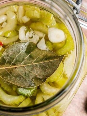 An open glass jar filled with pickled celery, sliced garlic, and a bay leaf, with a spoon containing pickled celery pieces beside the jar on a wooden surface.