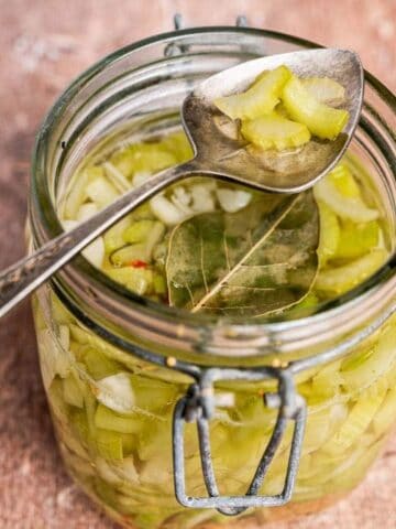 An open glass jar filled with pickled vegetables and a bay leaf, with a metal spoon resting on top, on a wooden surface.