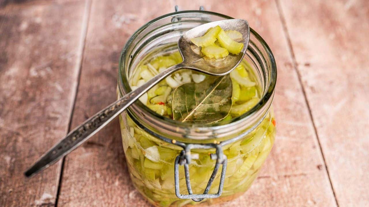 An open glass jar filled with pickled vegetables and a bay leaf, with a metal spoon resting on top, on a wooden surface.