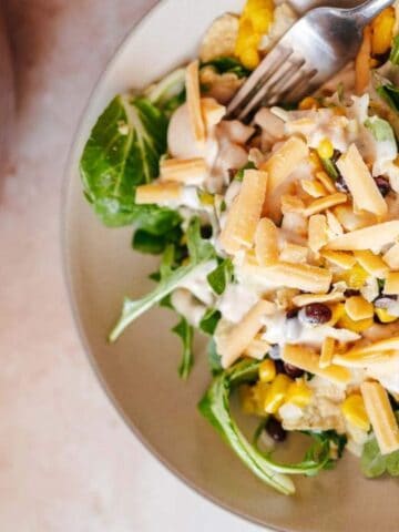 A bowl of salad with spinach, corn, black beans, shredded cheese, and creamy dressing, with a fork resting on the side.