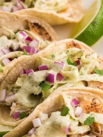 Three soft shell tacos filled with grilled chicken, diced red onion, chopped cilantro, shredded cabbage, and creamy sauce, served with lime wedges on a white rectangular plate.