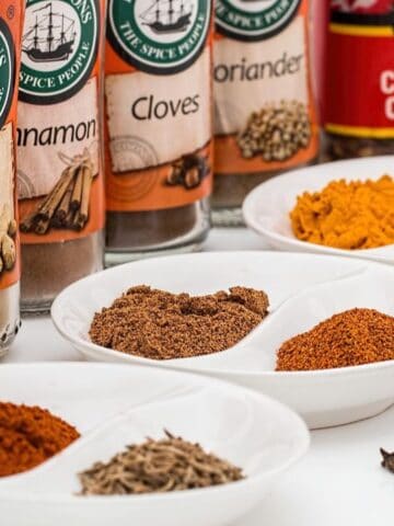 Assorted ground and whole spices are displayed in white dishes on a table, with spice jars labeled cumin, ginger, cinnamon, and more in the background.