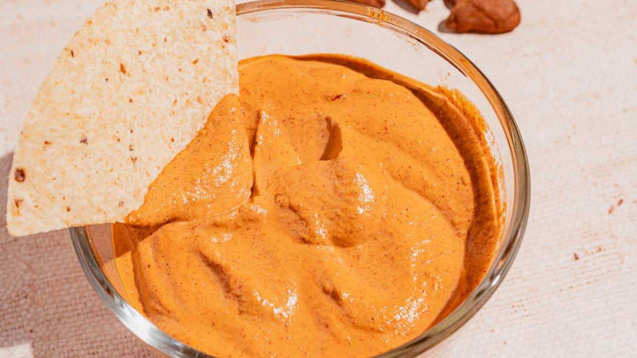 A glass bowl filled with creamy, orange-colored dip with a tortilla chip partially dipped into it.