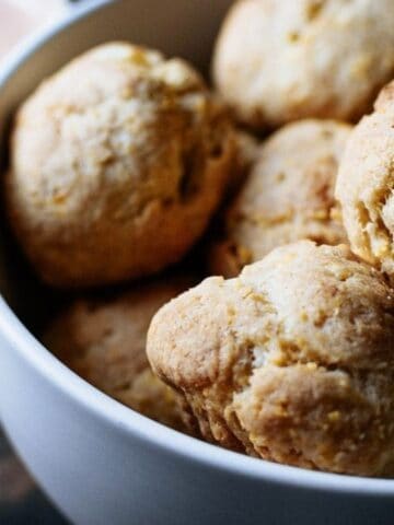 A white bowl filled with freshly baked biscuits sits next to a wooden spoon and a small dish of dipping sauce.