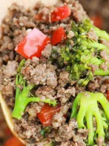 Close-up of cooked ground beef mixed with broccoli florets and chopped red bell peppers on a wooden spoon.