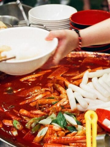 A person serves tteokbokki, Korean spicy rice cakes, from a large pan with red sauce, alongside bowls and skewered ingredients.