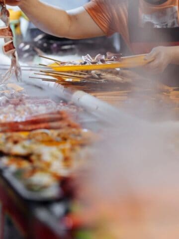 A food vendor prepares skewers of meat and seafood at a smoky market stall, handing a skewer to a customer.