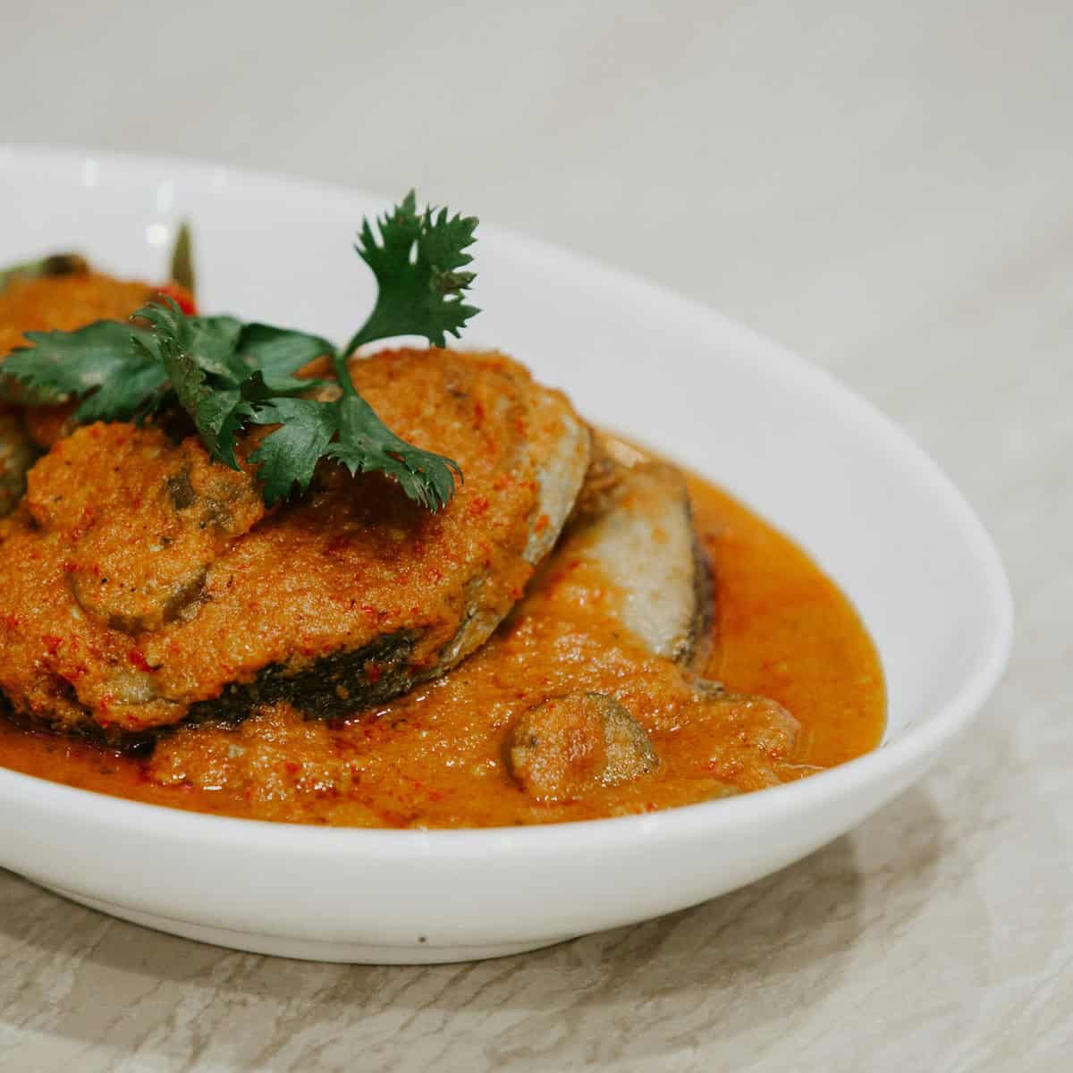 A white bowl containing fish in a thick orange curry sauce, garnished with a sprig of cilantro—classic Goan food at its finest.