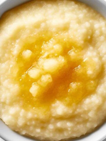 A white bowl filled with creamy grits, a classic among Bahamas breakfast foods, topped with melted butter and viewed from above on a white background.